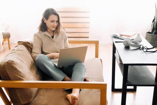 Joyful millennial woman typing on a laptop, attending a virtual meeting, seated on a sofa. Cheerful millennial female typing on laptop, has meeting with gadget, sit on sofa.