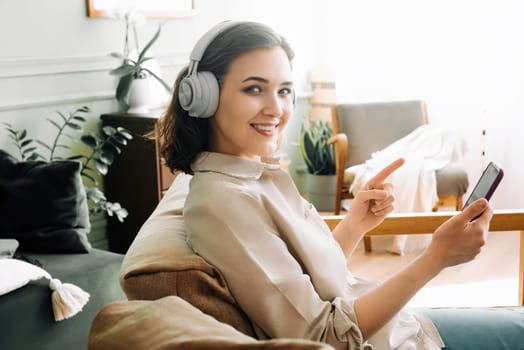 Happy Young Woman with Wireless Headphones Smiles, Points to Smartphone, Reading a Message. Joyful Young Woman with Wireless Headphones, Smiling, and Pointing at Smartphone While Reading a Message