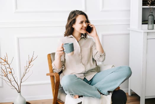 Cheerful Young Woman Talking on Smartphone, Relaxing in Armchair with Drink. Happy Young Woman Talking on Smartphone, Relaxing with Drink in Armchair.