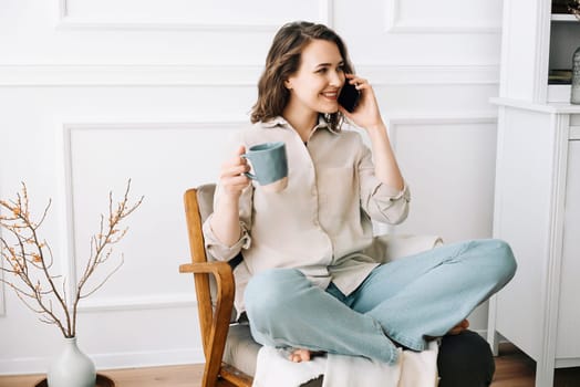 Cheerful Millennial Woman Engaging in Phone Call, Sipping Coffee, Sitting on Chair, Contemplating Empty Space.