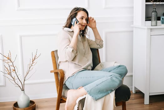 Serious millennial woman talks on the phone, sitting on a chair and looking out the window in her free time. Serious pretty millennial woman calls by phone, talks, sits on chair, looks out window.