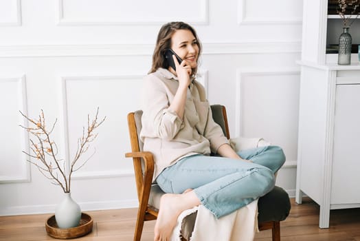 Millennial Woman Smiling, Speaking on Phone, Sitting on Chair, Looking Out Window at Free Space. Smiling millennial female calls by phone, talks, sits on chair looks out window at free space.