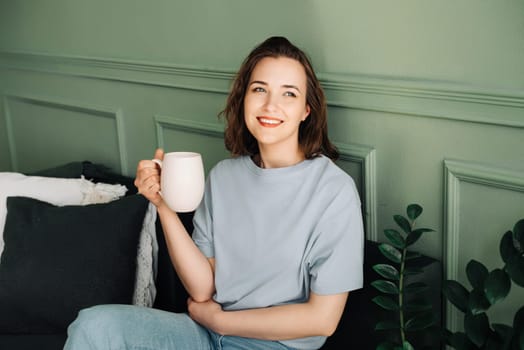 Cozy Tea Time - Cropped View of Woman Enjoying Tea on Living Room Couch. Relaxed Domestic Scene, Unwinding in Interior Ambiance. Female Hands Holding Mug with Tea or Coffee.