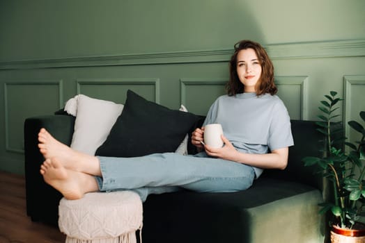 Contemplative Middle-Aged Woman Relaxing with Tea on Couch, Pensive, Gazing into Open Space. Reflective Moment: Thoughtful Woman Resting and Pondering on Sofa with Cup of Tea