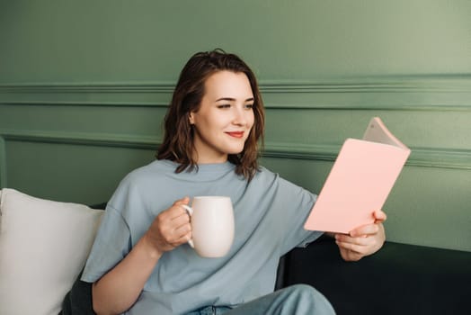 Relaxed Millennial Woman Reading Book, Enjoying Tea on Living Room Couch. Leisure and Relaxation. Joyful Young Woman Reading and Drinking Tea, Relaxing in Comfortable Home Environment