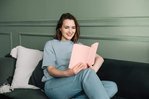 Happy Millennial Woman Enjoying Book on Cozy Living Room Couch. Relaxation and Leisure Time. Joyful Young Woman Reading Book in Comfortable Home Environment, Cozy Living Room Scene