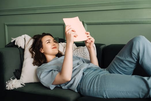 Contented Woman Relaxing on Sofa with Book, Enjoying Leisure Time. Smiling Young Lady Engaged in Reading While Lounging in Cozy Living Room Atmosphere. Relaxation and Enjoyment in Domestic Comfort