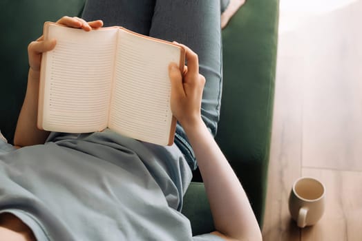 Relaxed Young Woman Enjoying Book, Tea on Living Room Sofa. Cozy Leisure Time. Lady Reading, Sipping Tea in Comfortable Home Setting. Relaxation and Gastronomic Pleasure in Domestic Atmosphere.