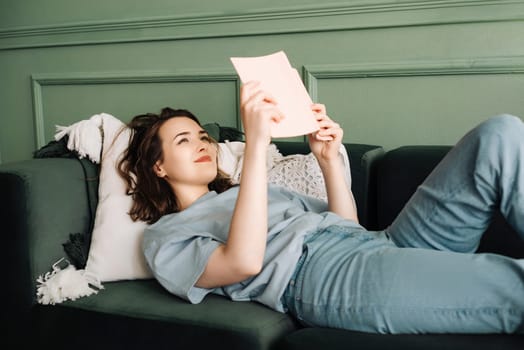 Relaxing Leisure. Smiling Woman Engrossed in Book, Lounging on Living Room Couch. Enjoying Quiet Reading Time at Home. Content and Relaxed Female Absorbed in Book on Cozy Sofa.