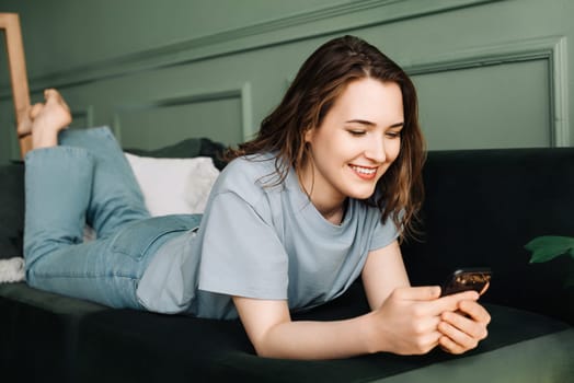 Sociable Relaxation. Woman Typing, Socializing on Smartphone While Lounging. Digital Socialization. Happy Woman Typing, Using Smartphone on Couch. Modern Relaxation.