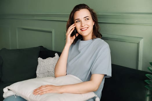 A smiling pretty young woman makes a call on her smartphone while enjoying a conversation in a gray living room. Joyful Chat. Pretty Woman Smiling during Smartphone Call.