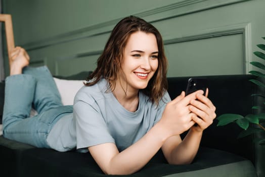 A cheerful young woman typing on her smartphone, using social media, socializing, lying on the couch. Connected and Content. Young Woman Engages on Smartphone, Relaxing on Couch.