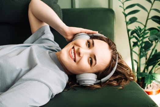 Joyful young woman wearing wireless headphones listening to music while lying on the sofa, enjoying her free time. Music Bliss. Happy Woman Relaxing on Sofa with Wireless Headphones.