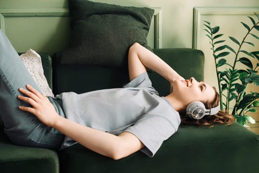 A smiling middle-aged woman wearing wireless headphones listens to music while lying on the sofa. Blissful Melodies. Middle-Aged Woman Enjoys Music with Wireless Headphones.