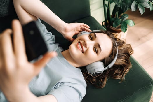 A smiling young middle-aged woman wearing wireless headphones listens to music while lying on the couch with her smartphone. Contentment in Tunes. Smiling Woman Enjoys Music with Wireless Headphones.