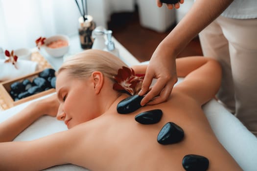 Hot stone massage at spa salon in luxury resort with day light serenity ambient, blissful woman customer enjoying spa basalt stone massage glide over body with soothing warmth. Quiescent