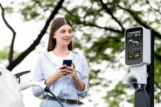 Young woman using smartphone online banking application to pay for electric car battery charging from EV charging station during vacation holiday road trip at national park or summer forest. Exalt