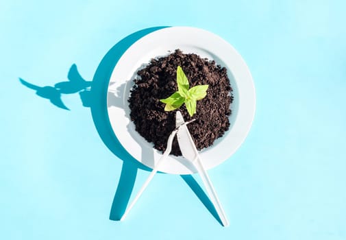 A plastic plate with earth and a green sprout, a fork crossed with a knife lie in the center on a light blue background with hard shadows, close-up flat lay. Ecology concept and restaurant etiquette sign.