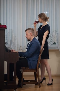 A pianist performs a piano piece from memory. Next to the man moves an elegantly dressed young woman. She drinks red wine from a glass.