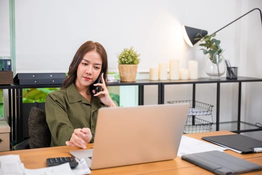 Happy Asian young woman working on laptop computer at home office while talking on mobile phone, smiling businesswoman having business call to talking with company sales client financial