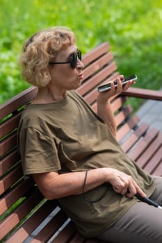 An elderly blind woman sits on a bench in the park and talks on a smartphone