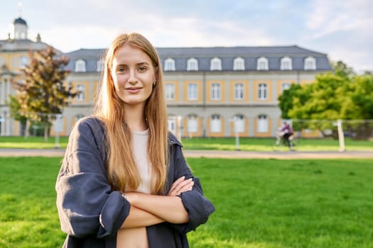 Portrait of young teenage girl student posing outdoors, background of educational building. Smiling female in glasses with textbooks standing on grass on lawn. Youth, education, college, university