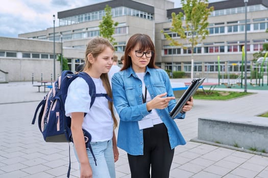 Talking female teacher and schoolgirl child outdoor, school building background. Meeting communication student girl with backpack and mentor counselor. Education, pre-teenage, learning, back to school