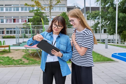 Talking female teacher and teenage high schoolgirl outdoor, school building background. Meeting communication student girl with backpack and mentor counselor. Education, adolescence, learning concept