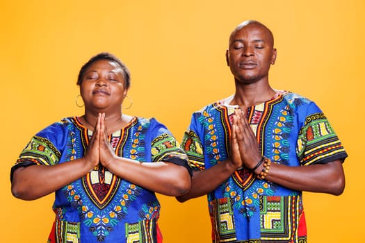 Black man and woman with closed eyes standing with folded palms, praying and asking for blessing. African american couple wearing ethnic clothes meditating and relaxing together