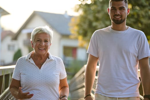 A handsome man and an older woman share a serene walk in nature, crossing a beautiful bridge against the backdrop of a stunning sunset, embodying the concept of a healthy and vibrant intergenerational life