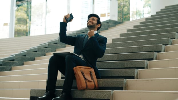 Successful business man celebrate successful project while sitting at stairs. Smart project manager getting new gob, getting promotion, increasing sales while calling friends by using phone. Exultant.