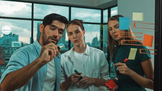 Professional business team brainstorm, sharing, thinking idea while skilled male leader write on sticky notes at glass wall. Skilled manager placed business plan on glass wall at office. Tracery
