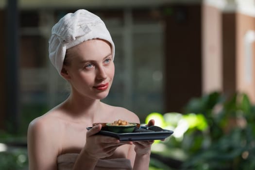 Portrait of beautiful caucasian woman in white towel holds the tray which contained the bowl of herbal scrub and spa rock surrounded by calm and relaxing nature. Side view. Tranquility.