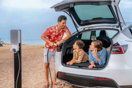 Family vacation trip traveling by the beach with electric car, lovely family sit on the trunk, charging EV car battery with green and sustainable energy. Family travel and eco-friendly car. Perpetual