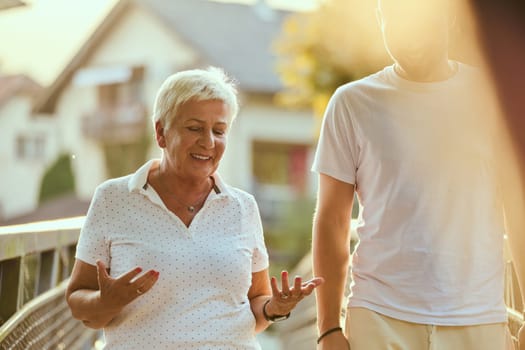 A handsome man and an older woman share a serene walk in nature, crossing a beautiful bridge against the backdrop of a stunning sunset, embodying the concept of a healthy and vibrant intergenerational life