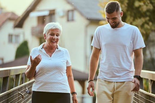 A handsome man and an older woman share a serene walk in nature, crossing a beautiful bridge against the backdrop of a stunning sunset, embodying the concept of a healthy and vibrant intergenerational life