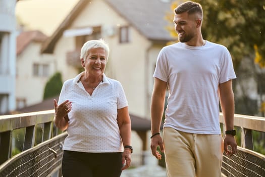 A handsome man and an older woman share a serene walk in nature, crossing a beautiful bridge against the backdrop of a stunning sunset, embodying the concept of a healthy and vibrant intergenerational life