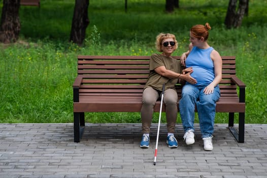 An elderly blind woman holds her pregnant daughter by the belly while sitting on a bench in the park