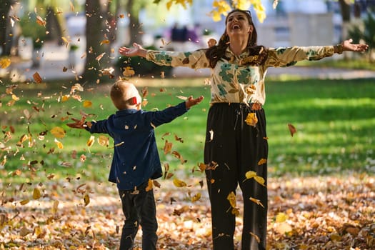 A modern woman joyfully plays with her son in the park, tossing leaves on a beautiful autumn day, capturing the essence of family life and the warmth of mother-son bonding in the midst of the fall season.