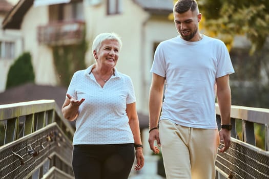 A handsome man and an older woman share a serene walk in nature, crossing a beautiful bridge against the backdrop of a stunning sunset, embodying the concept of a healthy and vibrant intergenerational life