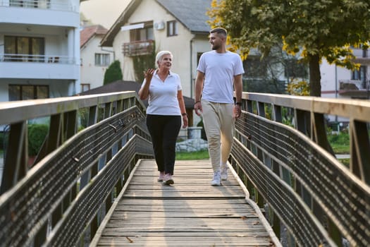 A handsome man and an older woman share a serene walk in nature, crossing a beautiful bridge against the backdrop of a stunning sunset, embodying the concept of a healthy and vibrant intergenerational life