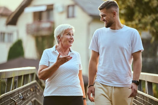 A handsome man and an older woman share a serene walk in nature, crossing a beautiful bridge against the backdrop of a stunning sunset, embodying the concept of a healthy and vibrant intergenerational life