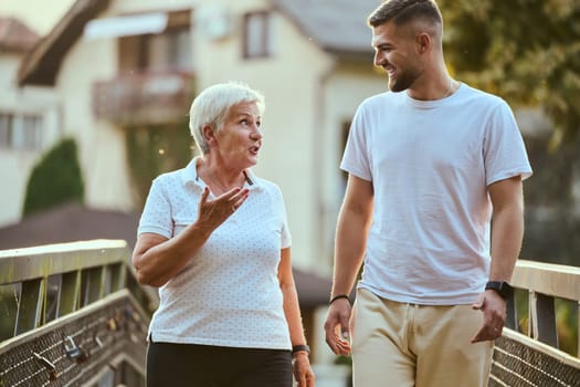 A handsome man and an older woman share a serene walk in nature, crossing a beautiful bridge against the backdrop of a stunning sunset, embodying the concept of a healthy and vibrant intergenerational life