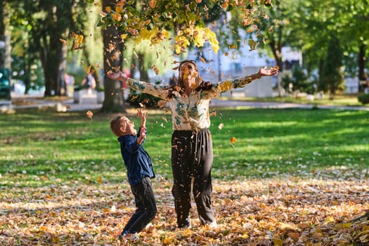 A modern woman joyfully plays with her son in the park, tossing leaves on a beautiful autumn day, capturing the essence of family life and the warmth of mother-son bonding in the midst of the fall season.