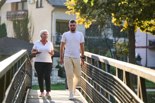 A handsome man and an older woman share a serene walk in nature, crossing a beautiful bridge against the backdrop of a stunning sunset, embodying the concept of a healthy and vibrant intergenerational life
