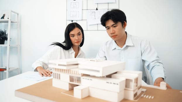 Closeup of skilled architecture engineer team measure and decide house model while working together and discussing about house structure at meeting room with blueprint placed behind. Immaculate.