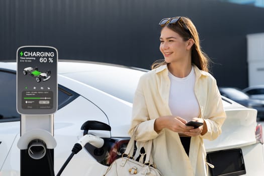 Young woman holding shopping bag and use smartphone to pay for electricity for recharging EV car battery from charging station at city mall parking lot. Modern woman go shopping by eco car. Expedient