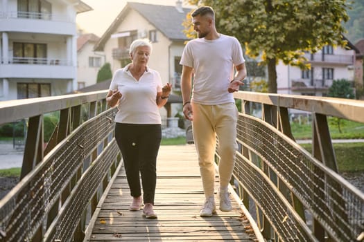 A handsome man and an older woman share a serene walk in nature, crossing a beautiful bridge against the backdrop of a stunning sunset, embodying the concept of a healthy and vibrant intergenerational life