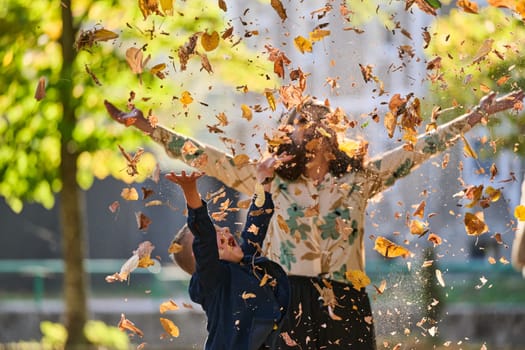 A modern woman joyfully plays with her son in the park, tossing leaves on a beautiful autumn day, capturing the essence of family life and the warmth of mother-son bonding in the midst of the fall season.