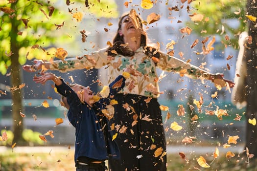 A modern woman joyfully plays with her son in the park, tossing leaves on a beautiful autumn day, capturing the essence of family life and the warmth of mother-son bonding in the midst of the fall season.